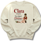 An off-white crewneck sweatshirt featuring a custom holiday graphic with the text 'Clara (noun): The original Christmas main character.' in red and black, accompanied by an illustration of a sleeping nutcracker and a gift box.