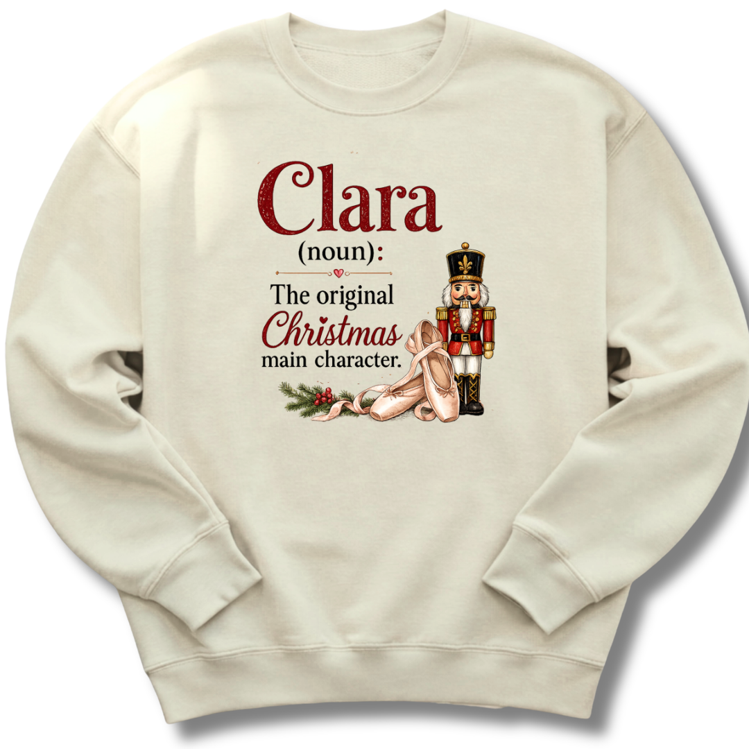 An off-white crewneck sweatshirt featuring a custom holiday graphic with the text 'Clara (noun): The original Christmas main character.' in red and black, accompanied by an illustration of a sleeping nutcracker and a gift box.