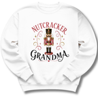A white crewneck sweatshirt featuring a graphic print of a nutcracker soldier with the words 'NUTCRACKER GRANDMA' in red and black lettering, surrounded by decorative gold swirls and stars.
