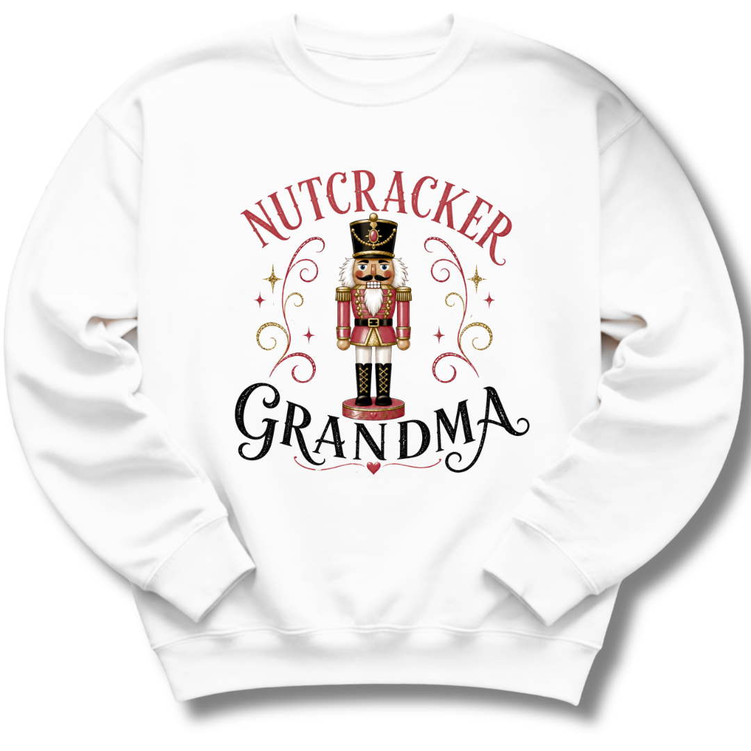 A white crewneck sweatshirt featuring a graphic print of a nutcracker soldier with the words 'NUTCRACKER GRANDMA' in red and black lettering, surrounded by decorative gold swirls and stars.
