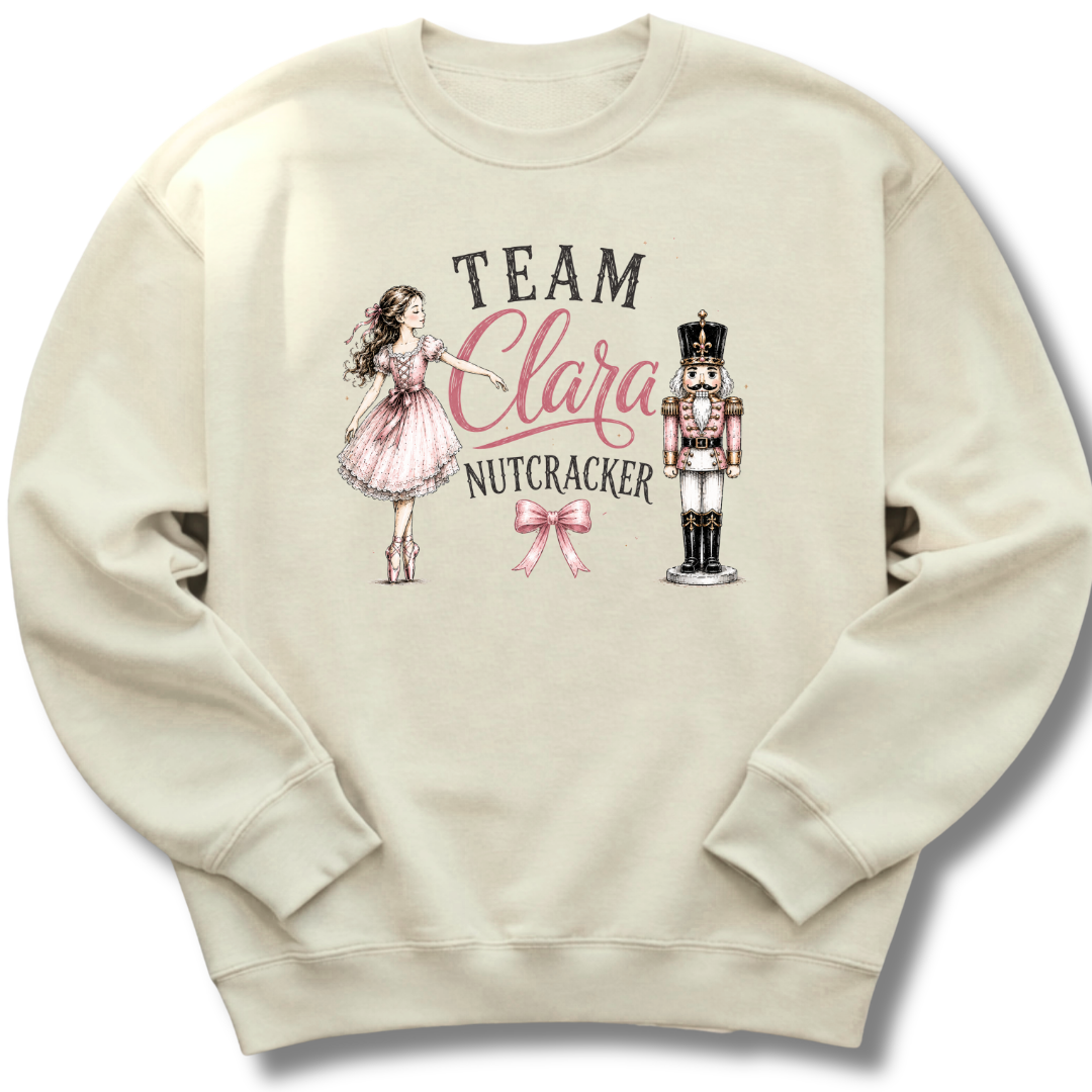 A cream-colored crewneck sweatshirt featuring a graphic print of a girl in a pink dress, a nutcracker, and the text 'TEAM Clara NUTCRACKER' with a pink bow.