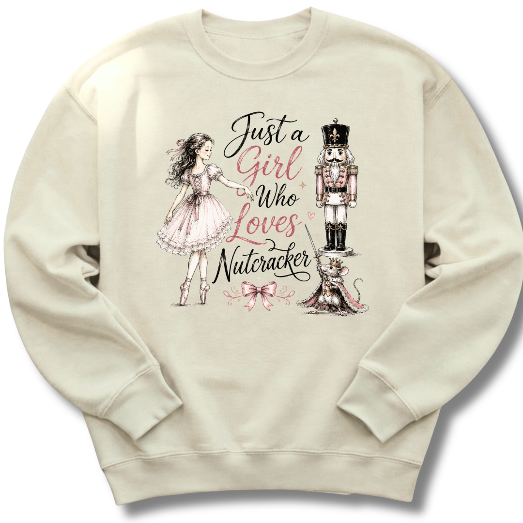A cream-colored crewneck sweatshirt featuring a graphic print of a ballerina, a nutcracker, a cat, and the text 'Just a Girl Who Loves Nutcracker' in black and pink lettering.