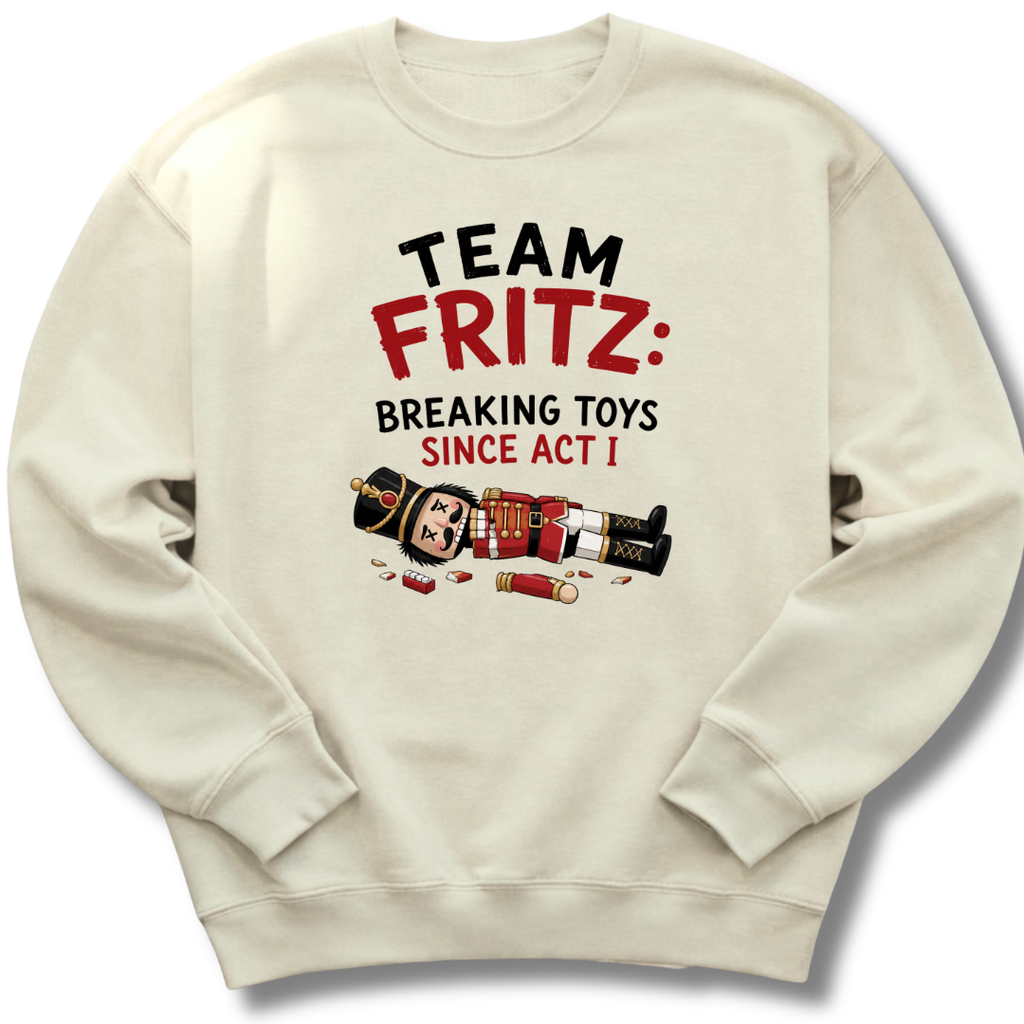 An off-white crewneck sweatshirt featuring a graphic print that reads 'TEAM FRITZ: BREAKING TOYS SINCE ACT I' in black and red text, accompanied by a cartoon illustration of a nutcracker and shotgun pieces scattered below.