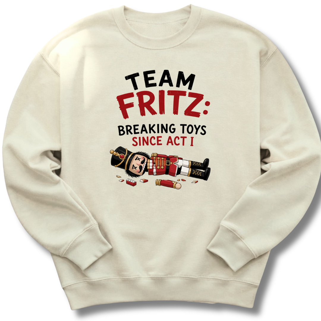 An off-white crewneck sweatshirt featuring a graphic print that reads 'TEAM FRITZ: BREAKING TOYS SINCE ACT I' in black and red text, accompanied by a cartoon illustration of a nutcracker and shotgun pieces scattered below.
