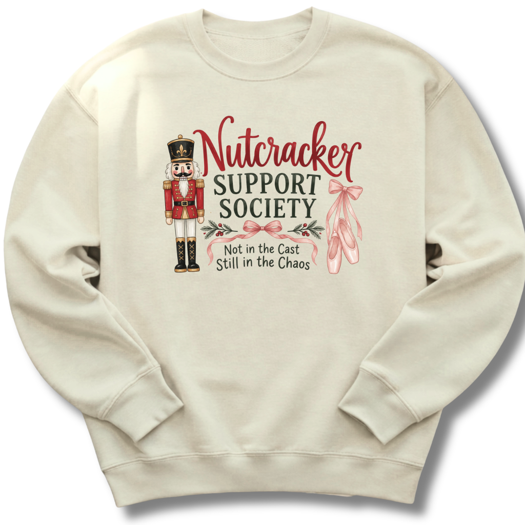 A cream-colored crewneck sweatshirt featuring a graphic design on the front. The design includes a nutcracker soldier, pink ballet slippers, and the text 'Nutcracker Support Society' and 'Not in the Cast Still in the Chaos'.