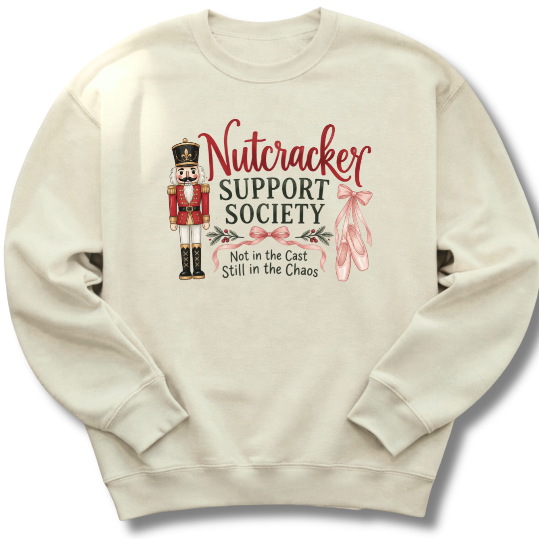 A cream-colored crewneck sweatshirt featuring a graphic design on the front. The design includes a nutcracker soldier, pink ballet slippers, and the text 'Nutcracker Support Society' and 'Not in the Cast Still in the Chaos'.