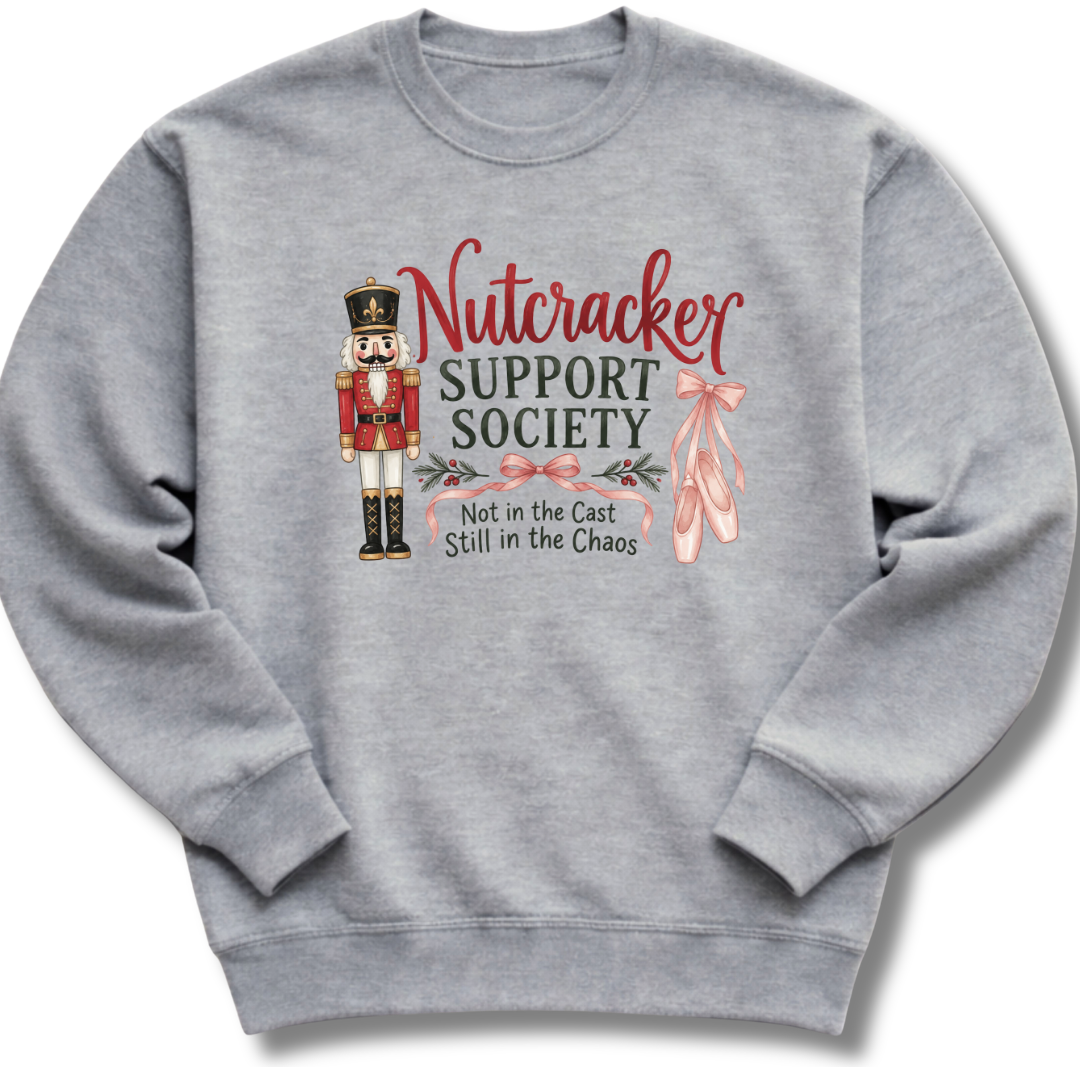 A heather gray crewneck sweatshirt featuring a Nutcracker-themed graphic with the text 'Nutcracker Support Society' and 'Not in the Cast Still in the Chaos' alongside a nutcracker figure, pink bows, and ballet slippers.