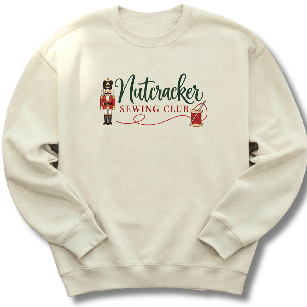 An off-white crewneck sweatshirt featuring a 'Nutcracker Sewing Club' graphic on the chest with a nutcracker figure, green script text, and red text.