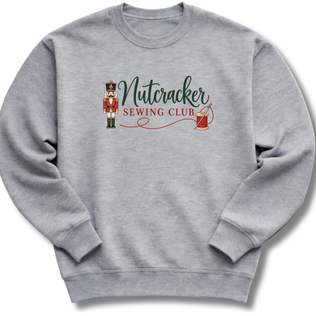 A heather gray crewneck sweatshirt featuring a 'Nutcracker Sewing Club' graphic on the chest with a nutcracker figure, green script text, and red text and line details.