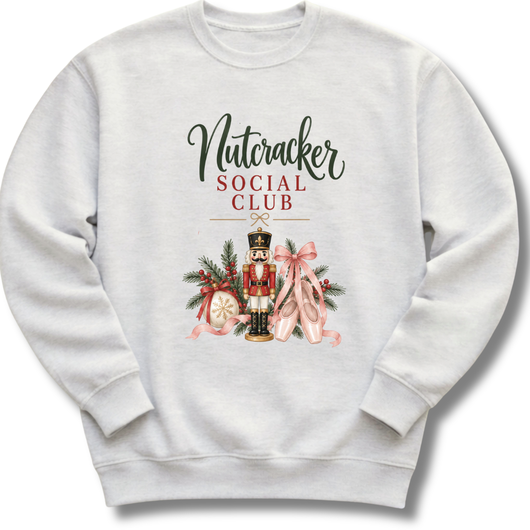 A light gray crewneck sweatshirt featuring a festive graphic print with the text 'Nutcracker Social Club' above an illustration of a nutcracker, pink ballet slippers, pine branches, and a red bow.