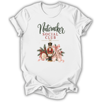 A white short-sleeve crewneck t-shirt with a graphic print on the front. The graphic features the text 'Nutcracker SOCIAL CLUB' in black and red lettering above an illustration of a nutcracker soldier decorated with a pink bow, green pine branches, and a gold hat.