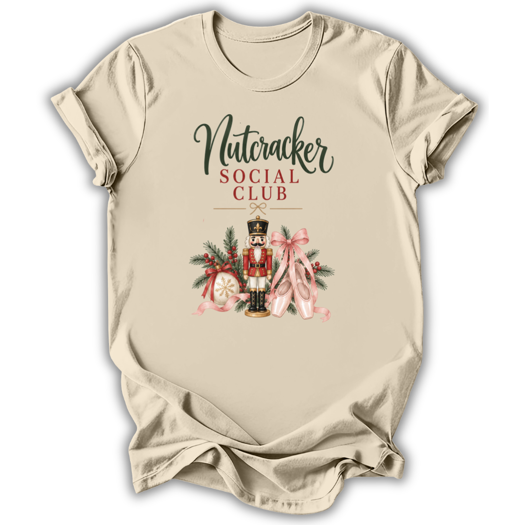 A beige short-sleeve t-shirt with a 'Nutcracker Social Club' graphic on the front, featuring a nutcracker, pink ribbons, and holiday greenery, displayed on a white background.
