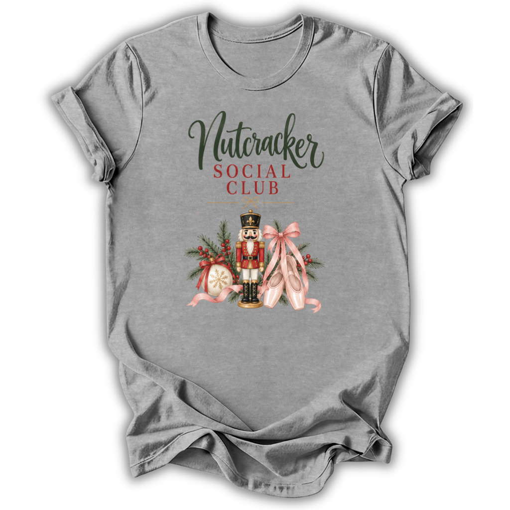 A heather gray short-sleeve t-shirt with a graphic print on the front reading 'Nutcracker Social Club' in dark green and red lettering, featuring a nutcracker, pink ribbons, and holiday ornaments.