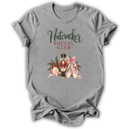 A heather gray short-sleeve t-shirt with a graphic print on the front reading 'Nutcracker Social Club' in dark green and red lettering, featuring a nutcracker, pink ribbons, and holiday ornaments.