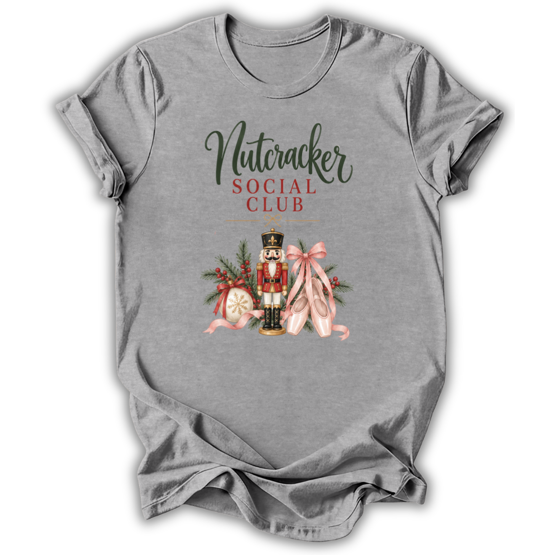 A heather gray short-sleeve t-shirt with a graphic print on the front reading 'Nutcracker Social Club' in dark green and red lettering, featuring a nutcracker, pink ribbons, and holiday ornaments.