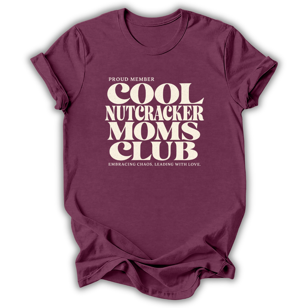 A maroon short-sleeve crewneck t-shirt with a cream-colored retro graphic that reads 'PROUD MEMBER COOL NUTCRACKER MOMS CLUB' and 'EMBRACING CHANGE, LEADING WITH LOVE.'.