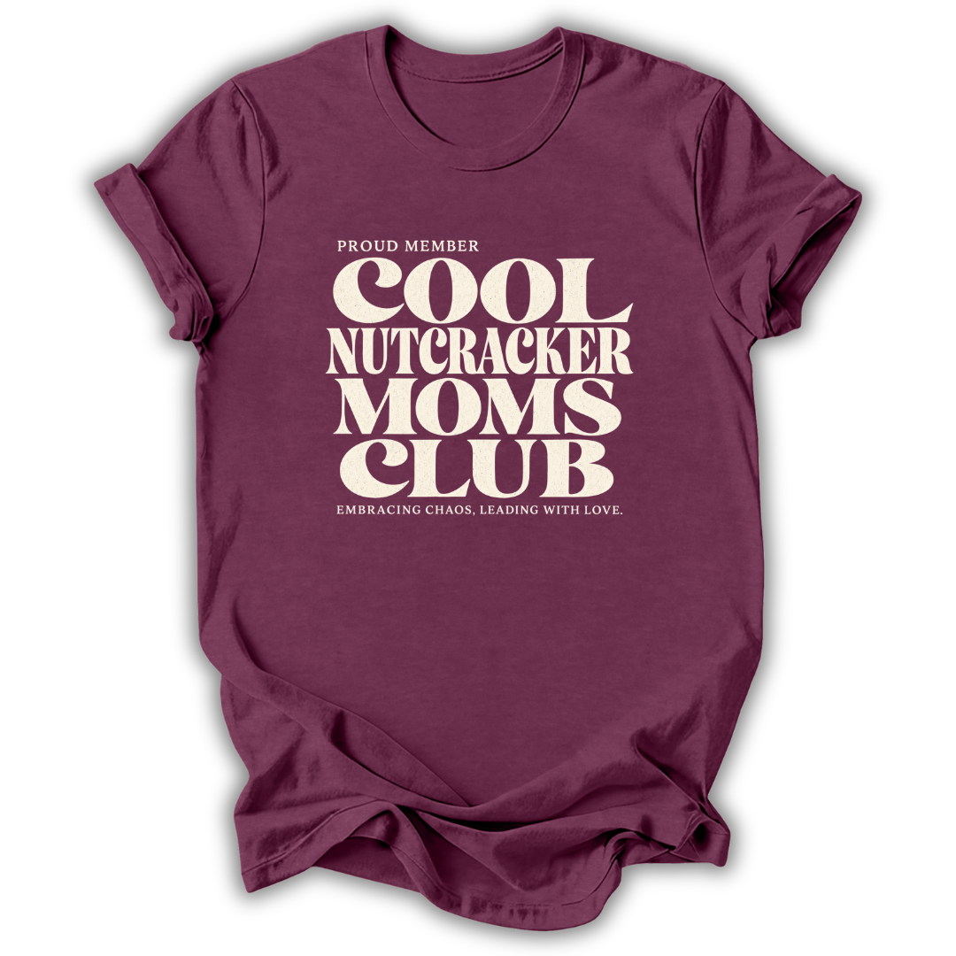 A maroon short-sleeve crewneck t-shirt with a cream-colored retro graphic that reads 'PROUD MEMBER COOL NUTCRACKER MOMS CLUB' and 'EMBRACING CHANGE, LEADING WITH LOVE.'.