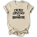A beige short-sleeve t-shirt with a black graphic text print reading 'I'M NOT OBSESSED I'M SUPPORTIVE' on the front, displayed against a white background with a soft drop shadow.