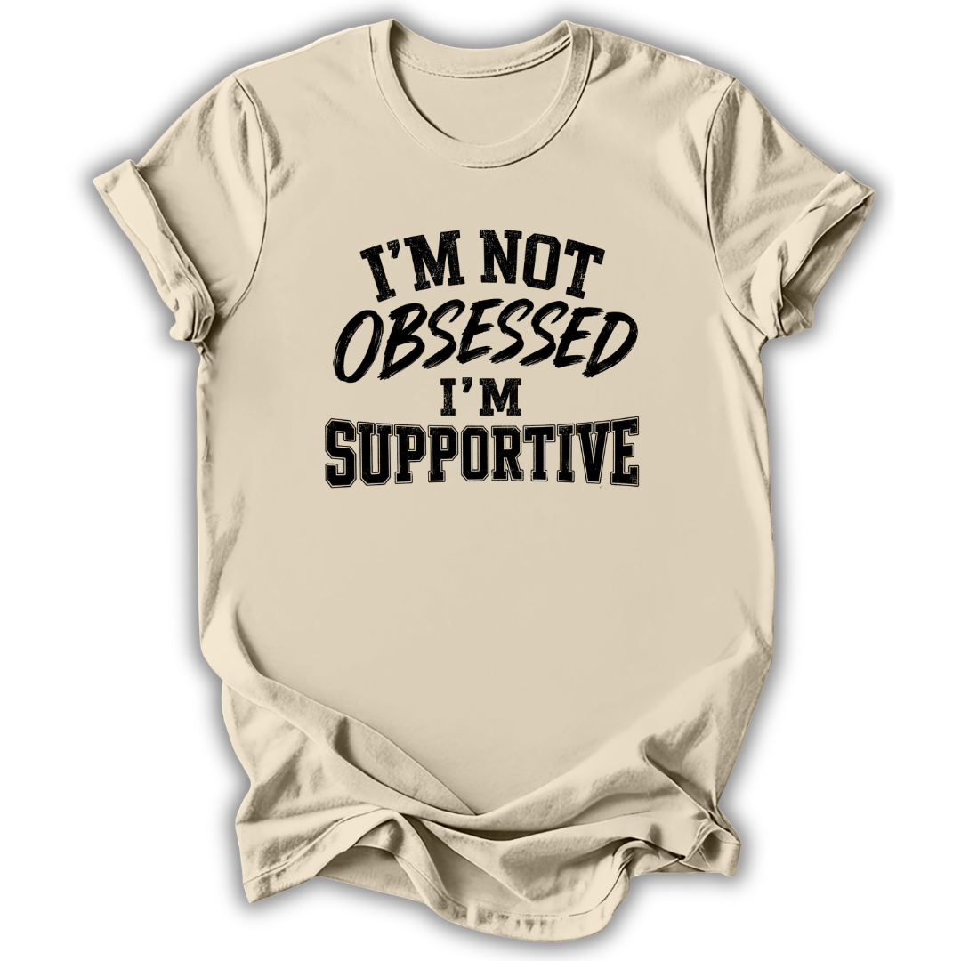 A beige short-sleeve t-shirt with a black graphic text print reading 'I'M NOT OBSESSED I'M SUPPORTIVE' on the front, displayed against a white background with a soft drop shadow.
