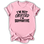 A light pink short-sleeve crewneck t-shirt with the text 'I'M NOT OBSESSED I'M SUPPORTIVE' printed in black across the chest, shown on a white background with a drop shadow.