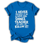 A bright blue short-sleeve t-shirt with white text that reads 'I NEVER DREAMED I'D BE A SUPER COOL DANCE TEACHER BUT HERE I AM KILLIN' IT!' displayed on a clean white background.