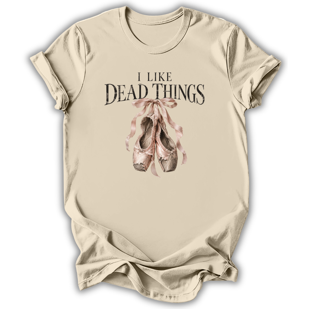 A beige short-sleeve t-shirt with a graphic print that reads 'I LIKE DEAD THINGS' above a pair of dusty pink and brown ballet slippers tied with a ribbon, shown on a white background.