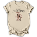 A beige short-sleeve t-shirt with a graphic print that reads 'I LIKE DEAD THINGS' above a pair of dusty pink and brown ballet slippers tied with a ribbon, shown on a white background.