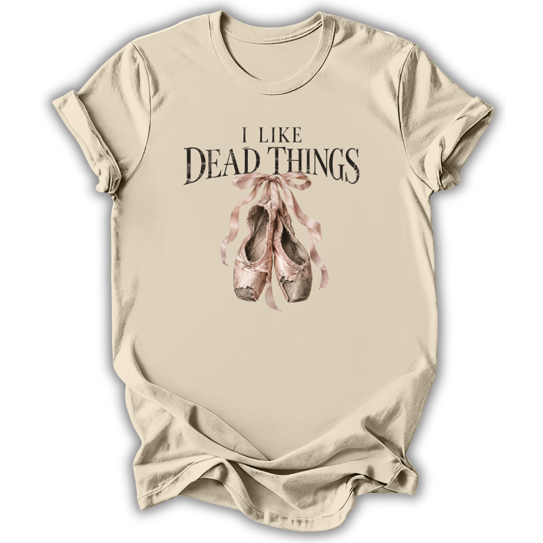 A beige short-sleeve t-shirt with a graphic print that reads 'I LIKE DEAD THINGS' above a pair of dusty pink and brown ballet slippers tied with a ribbon, shown on a white background.