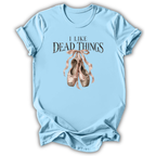 A light blue short-sleeve t-shirt with a graphic print that reads 'I LIKE DEAD THINGS' above a pair of dusty pink ballet slippers tied with pink ribbons, shown on a white background.