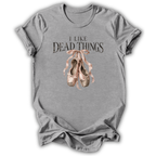 A heather gray short-sleeve t-shirt with a graphic print that reads 'I LIKE DEAD THINGS' above a pair of dusty pink ballet pointe shoes, shown on a white background.