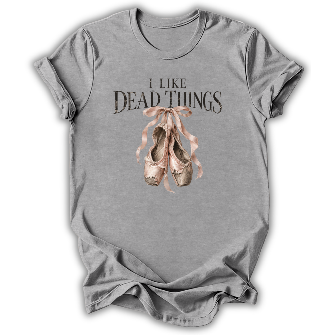 A heather gray short-sleeve t-shirt with a graphic print that reads 'I LIKE DEAD THINGS' above a pair of dusty pink ballet pointe shoes, shown on a white background.