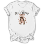 A white short-sleeve crewneck t-shirt with a graphic print on the front. The print features the text 'I LIKE DEAD THINGS' in a dark, gothic font above a watercolor illustration of dusty pink and brown ballet pointe shoes tied with a ribbon.