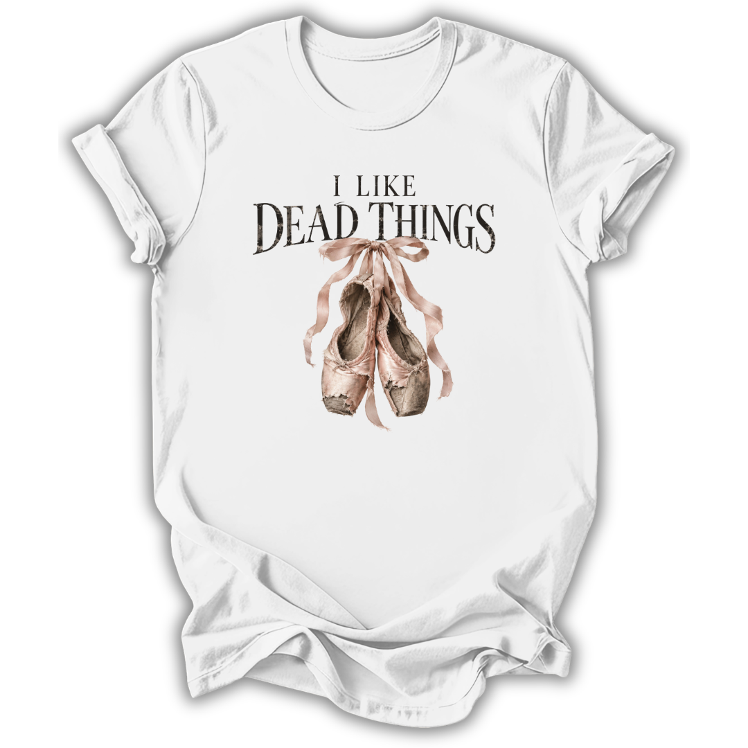 A white short-sleeve crewneck t-shirt with a graphic print on the front. The print features the text 'I LIKE DEAD THINGS' in a dark, gothic font above a watercolor illustration of dusty pink and brown ballet pointe shoes tied with a ribbon.