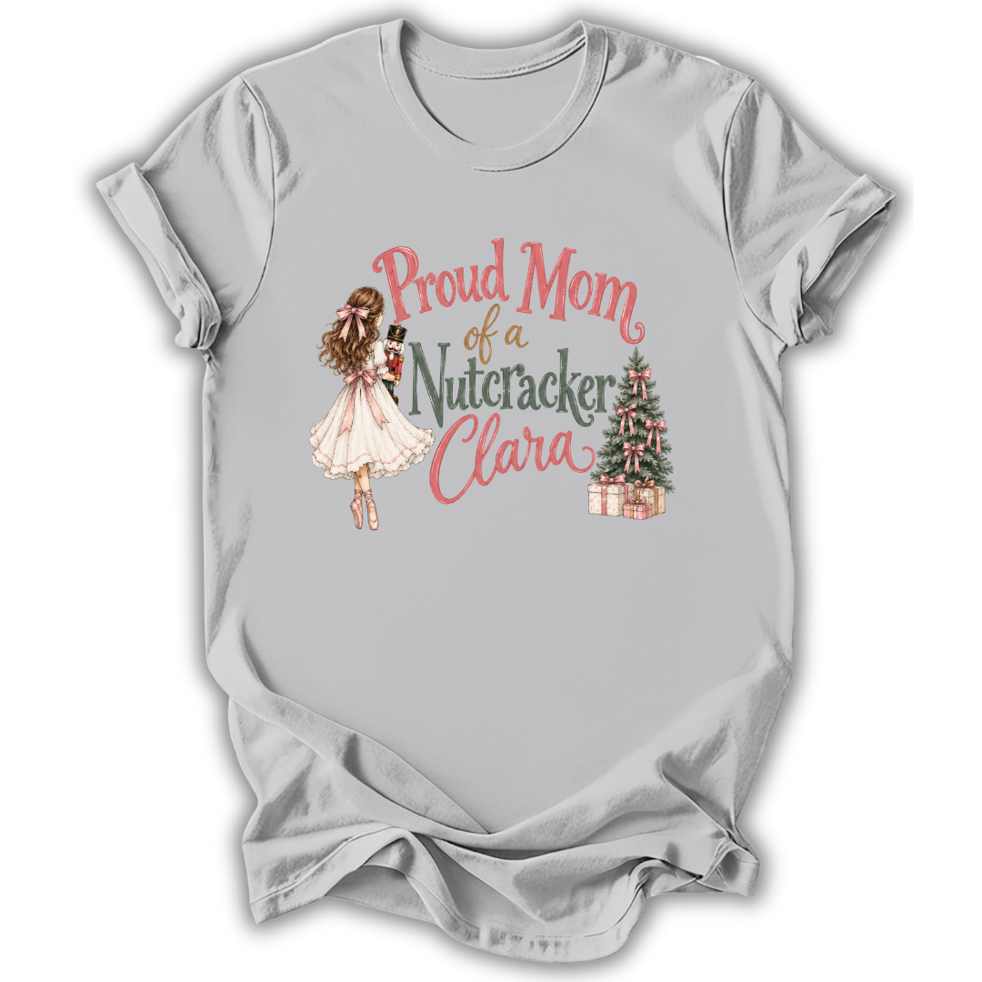 A light gray short-sleeve t-shirt with a graphic print on the front. The graphic features a vintage-style illustration of a woman in a white dress holding a nutcracker, next to the text 'Proud Mom of a Nutcracker Clara' in pink and green lettering, and a small decorated Christmas tree with wrapped gifts.