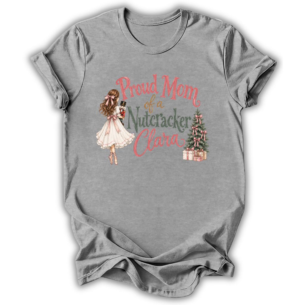 A heather gray short-sleeve t-shirt with a graphic print on the front. The print features an illustration of a woman in a white dress holding a nutcracker, next to the text 'Proud Mom of a Nutcracker Clara' in coral and green lettering, with a small decorated Christmas tree and wrapped gifts.