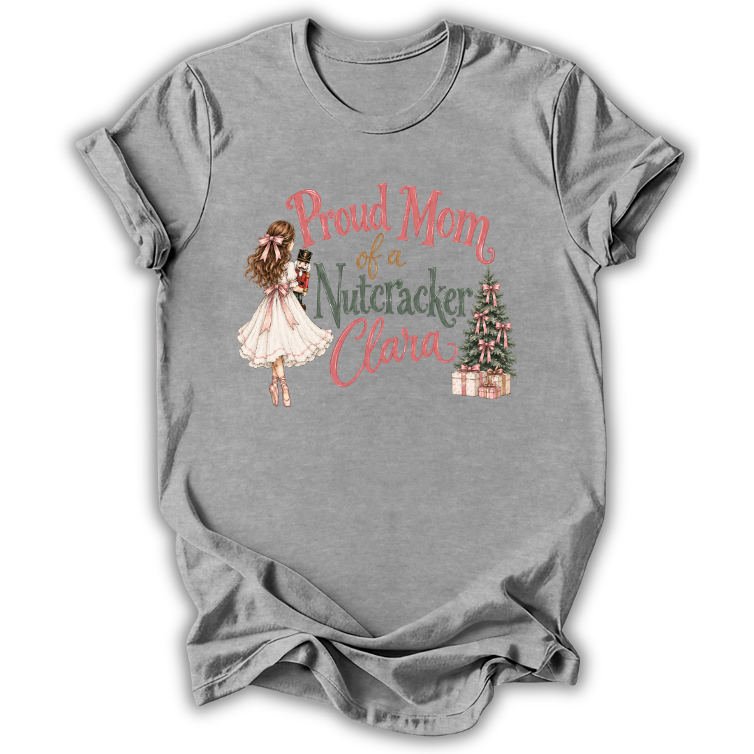 A heather gray short-sleeve t-shirt with a graphic print on the front. The print features an illustration of a woman in a white dress holding a nutcracker, next to the text 'Proud Mom of a Nutcracker Clara' in coral and green lettering, with a small decorated Christmas tree and wrapped gifts.
