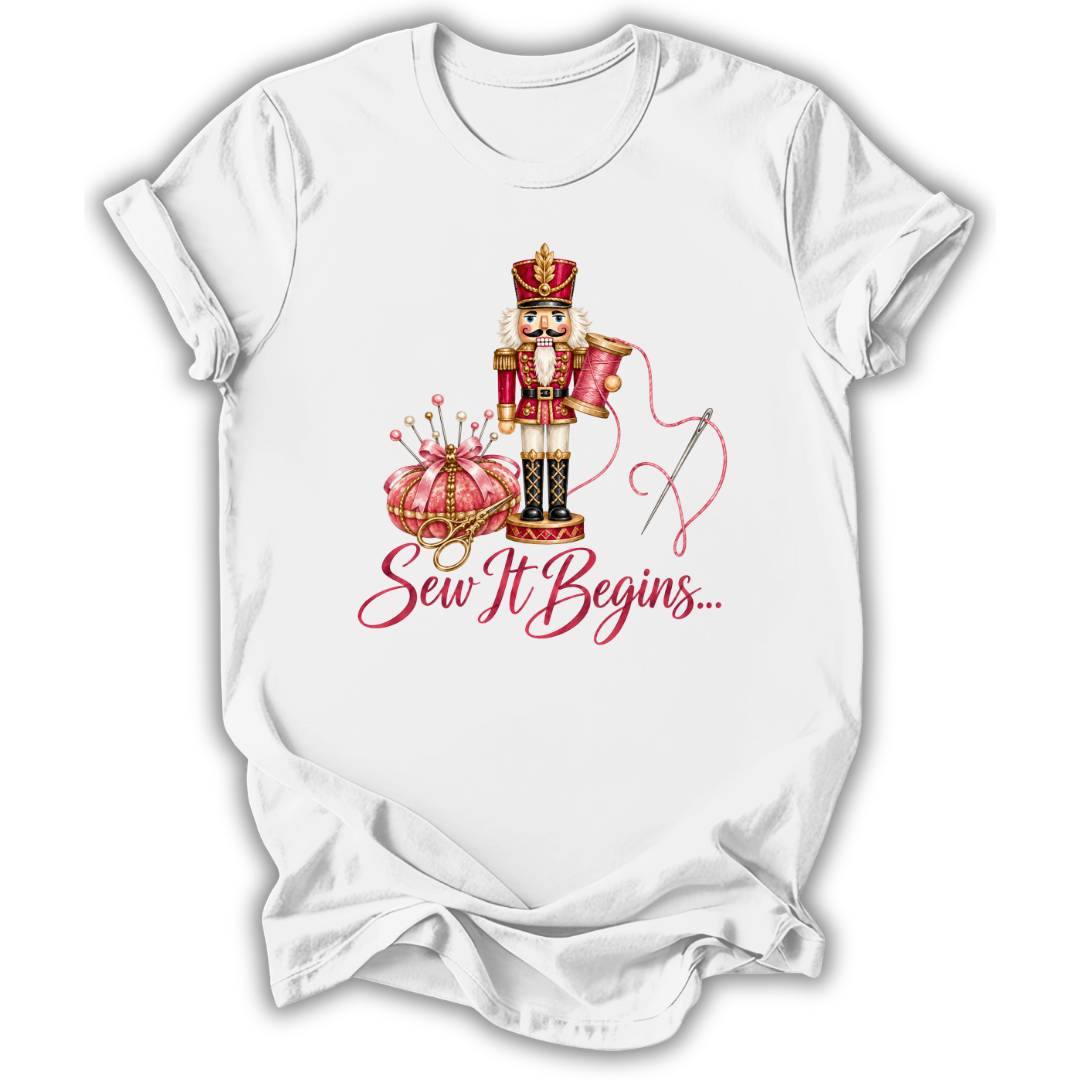 A white short-sleeve crewneck t-shirt with a graphic print of a nutcracker, a pink pumpkin, a needle, and the text 'Sew It Begins.