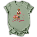 A sage green short-sleeve t-shirt with a graphic of a nutcracker, a pink pumpkin, and the text 'Sew It Begins.