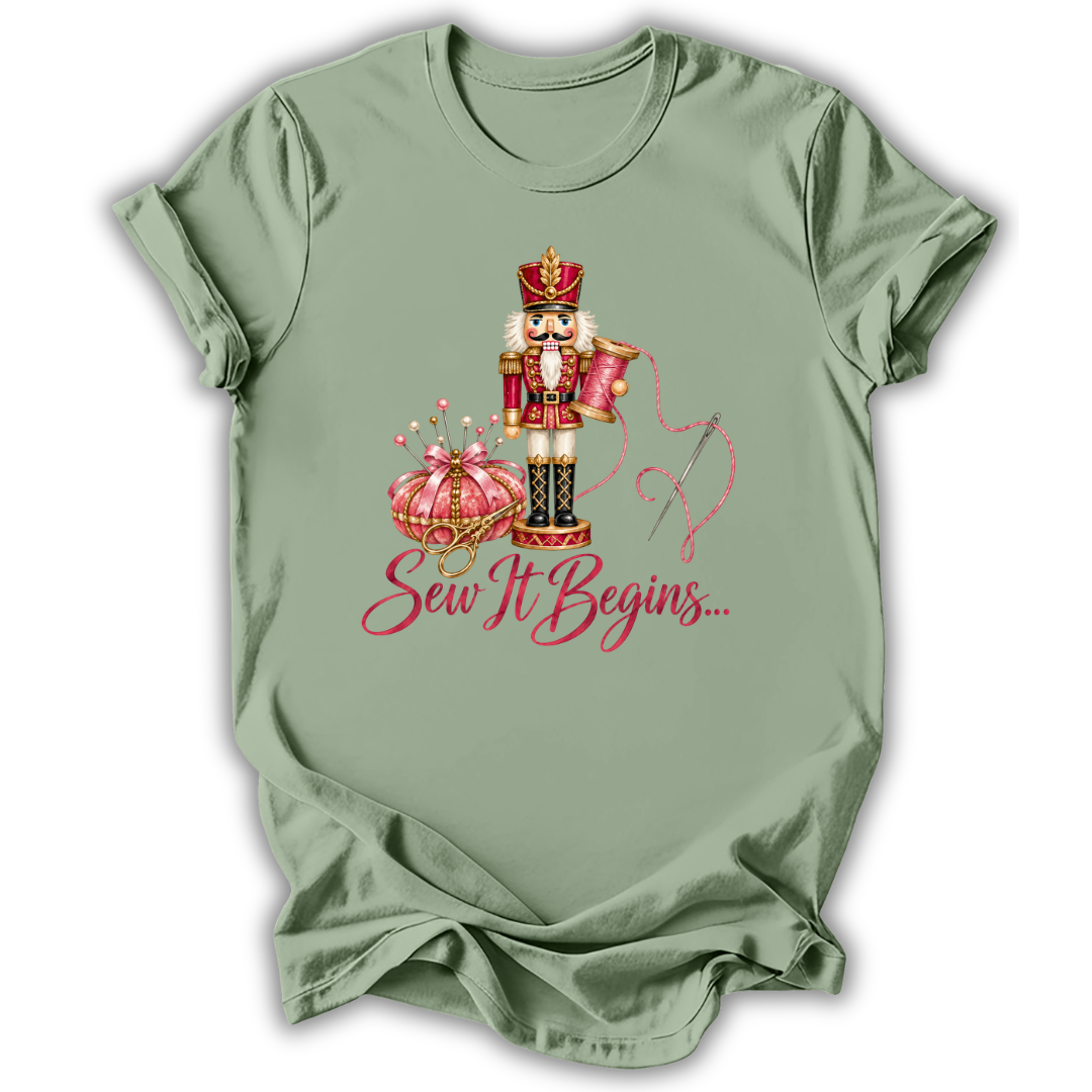 A sage green short-sleeve t-shirt with a graphic of a nutcracker, a pink pumpkin, and the text 'Sew It Begins.