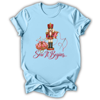 A light blue short-sleeve t-shirt with a graphic of a nutcracker, a pink pumpkin, a needle, and the text 'Sew It Begins.
