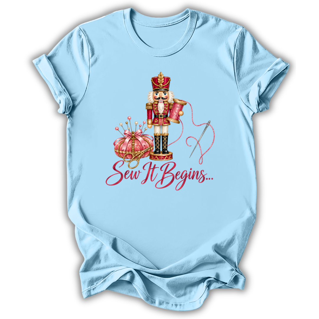 A light blue short-sleeve t-shirt with a graphic of a nutcracker, a pink pumpkin, a needle, and the text 'Sew It Begins.
