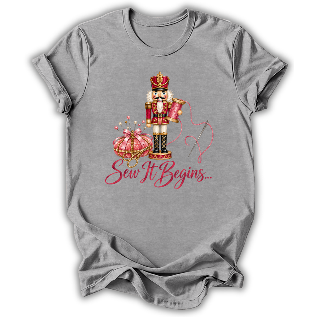 A heather gray short-sleeve t-shirt with a graphic of a nutcracker, a pink pumpkin, and the text 'Sew It Begins.