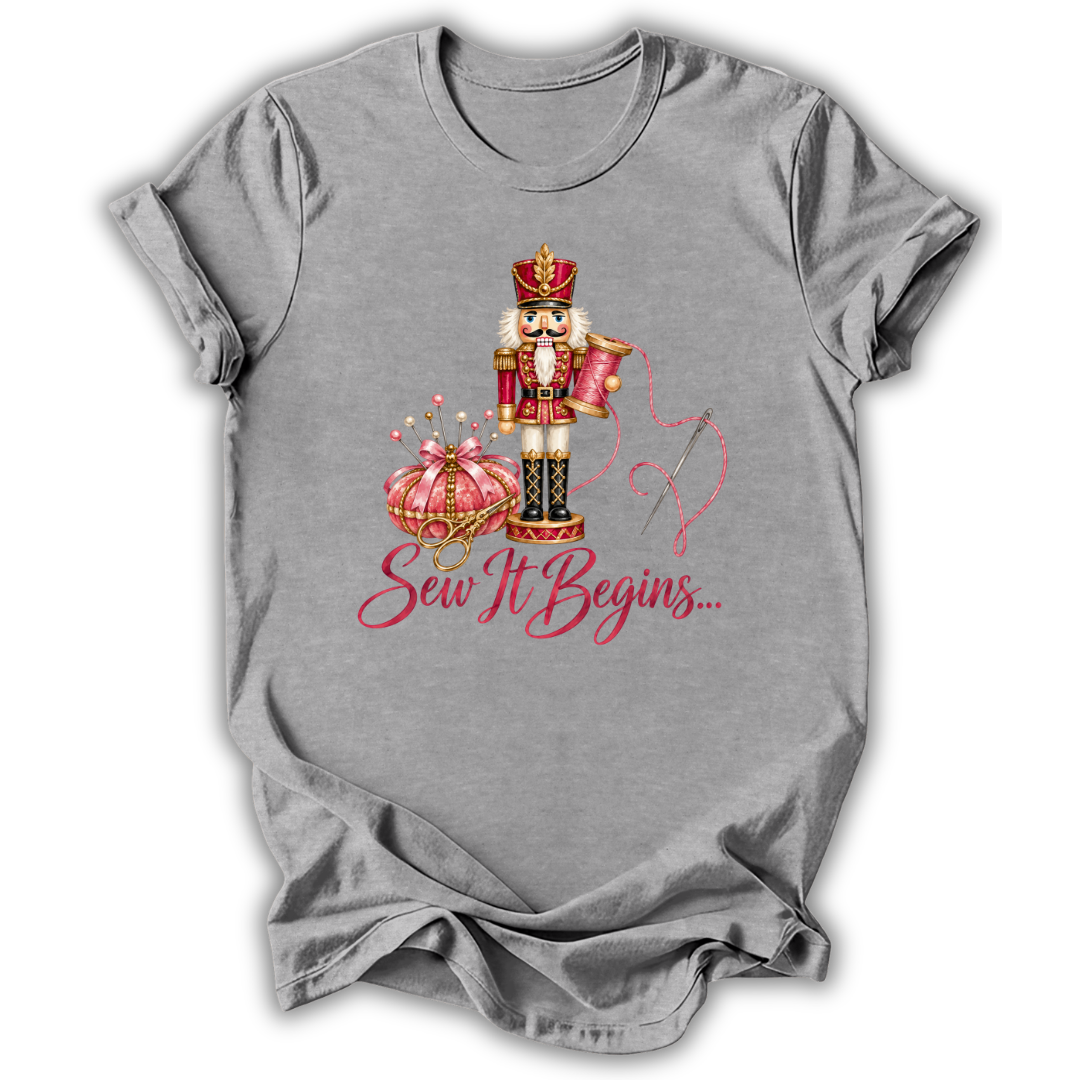 A heather gray short-sleeve t-shirt with a graphic of a nutcracker, a pink pumpkin, and the text 'Sew It Begins.