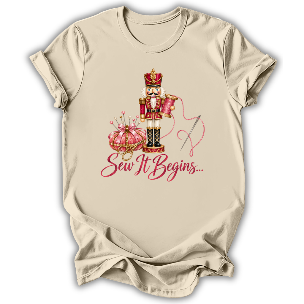 A beige short-sleeve crewneck t-shirt with a graphic of a nutcracker, a pink pumpkin, a needle, and the text 'Sew It Begins.