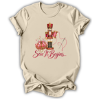 A beige short-sleeve crewneck t-shirt with a graphic of a nutcracker, a pink pumpkin, a needle, and the text 'Sew It Begins.