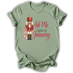 An olive green short-sleeve t-shirt with a graphic of a nutcracker holding a pink bow, next to the text 'Ask Me Again in January' in pink and brown lettering.