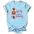 A light blue short-sleeve t-shirt with a graphic of a nutcracker holding pink slippers next to the text 'Ask Me Again in January' in red and dark green lettering, displayed on a clean white background.