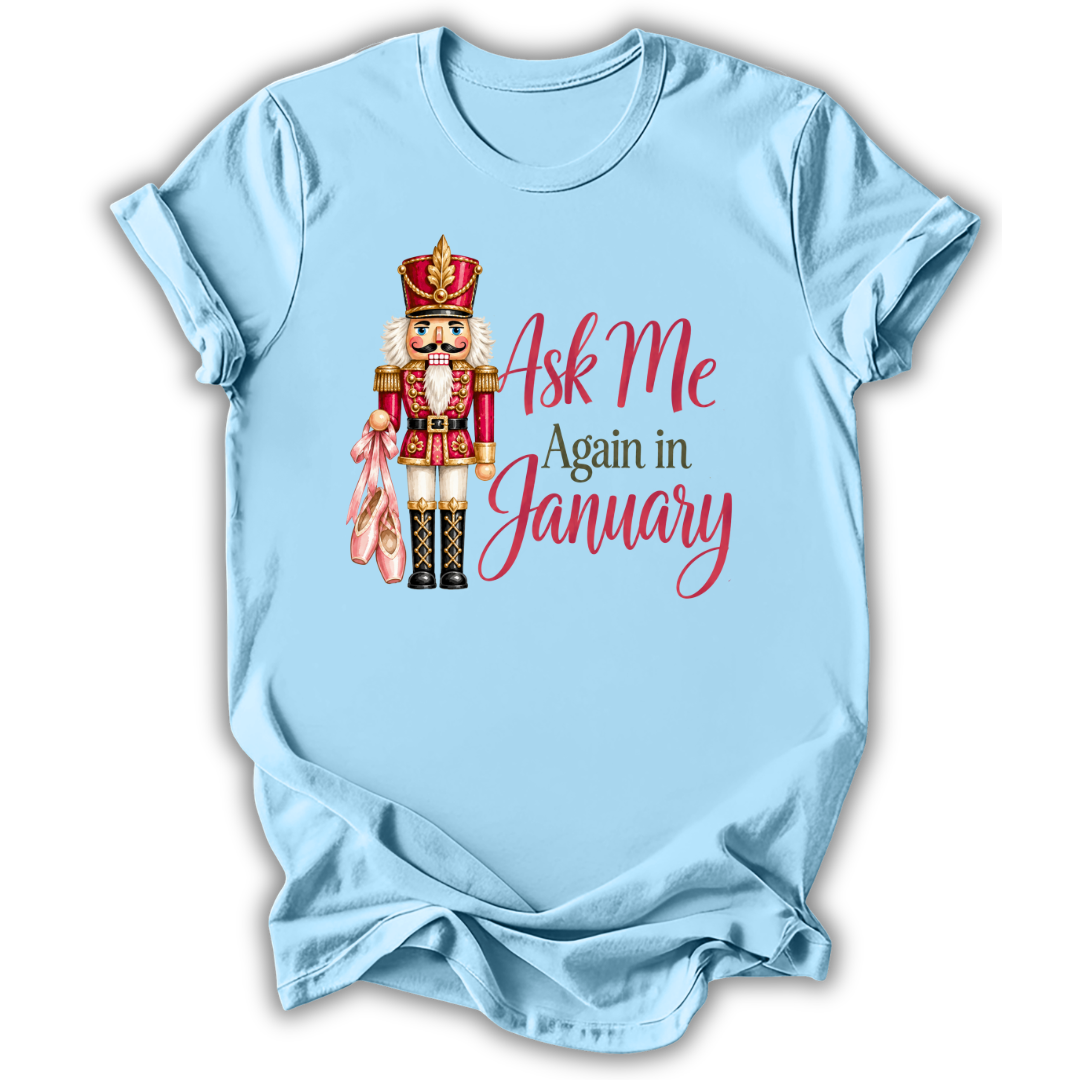 A light blue short-sleeve t-shirt with a graphic of a nutcracker holding pink slippers next to the text 'Ask Me Again in January' in red and dark green lettering, displayed on a clean white background.