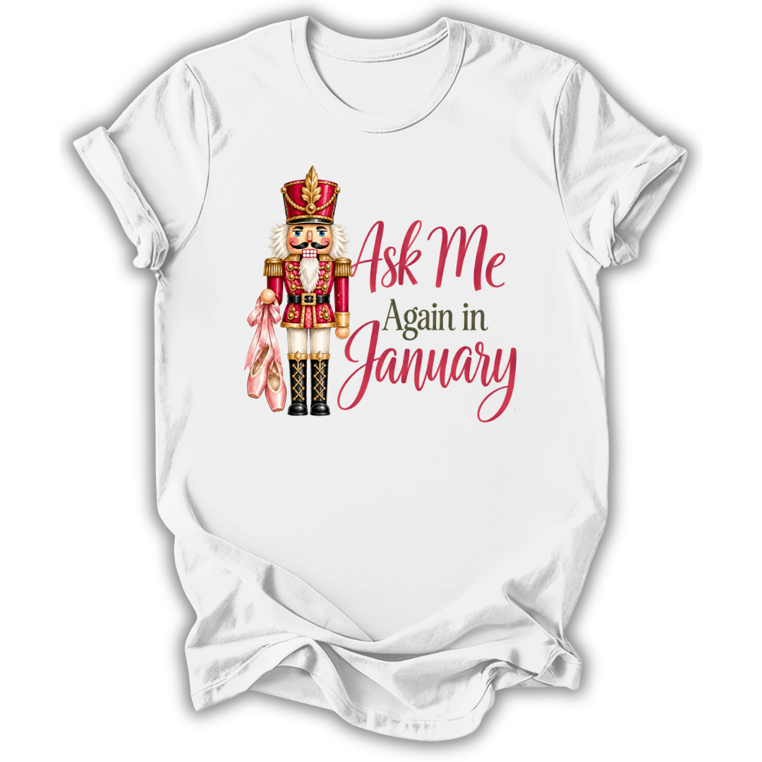 A white short-sleeve crewneck t-shirt with a graphic of a nutcracker holding a pink bow, next to the text 'Ask Me Again in January' in pink and dark green lettering.