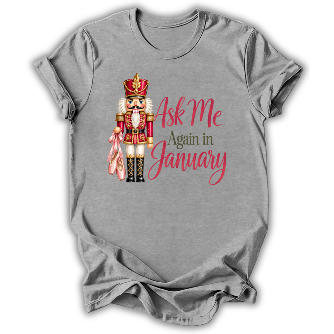 A heather gray short-sleeve t-shirt with a graphic of a nutcracker holding a pink bow and the text 'Ask Me Again in January' in pink and brown lettering.